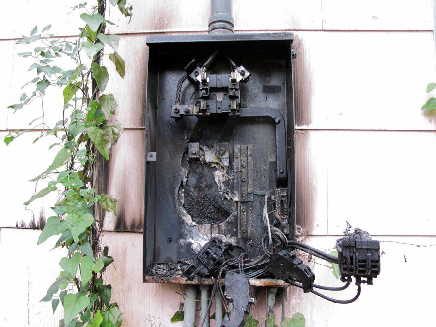 Burnt electrical panel on a building exterior, showing severe fire damage and charred components.