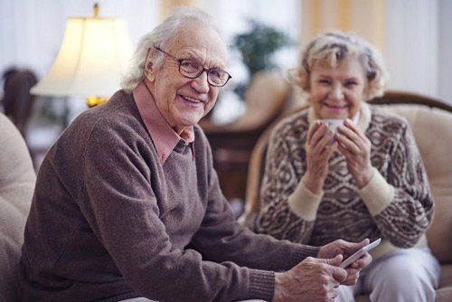 An elderly couple is sitting on a couch drinking coffee and looking at a cell phone.