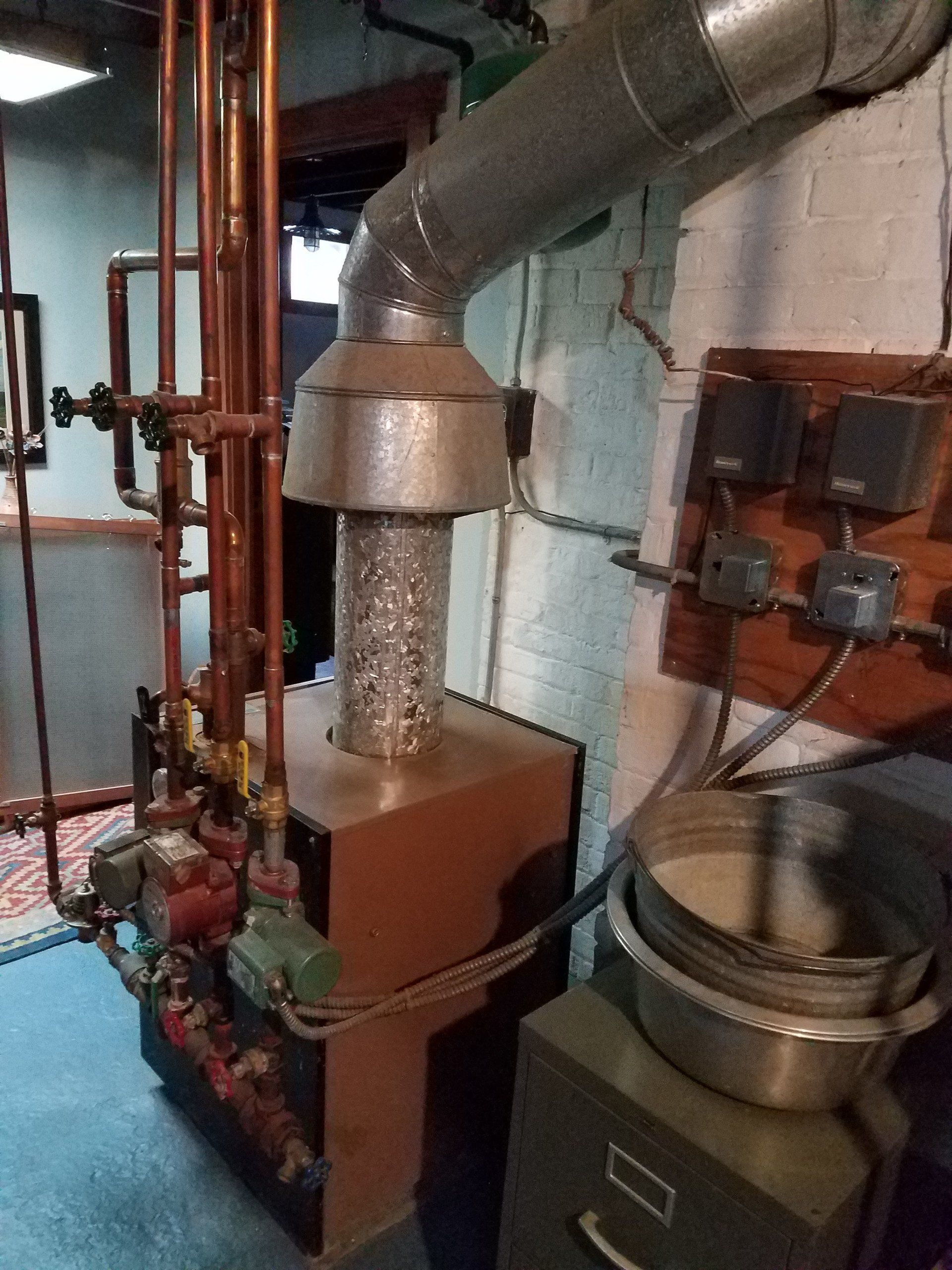 Boiler room with metal furnace, ductwork, pipes, and gauges against a brick wall.