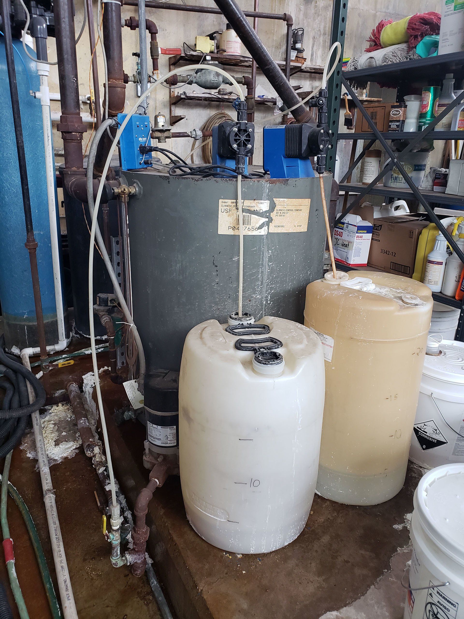 Industrial utility room with large water tanks, pipes, and equipment cluttered on shelving and the floor
