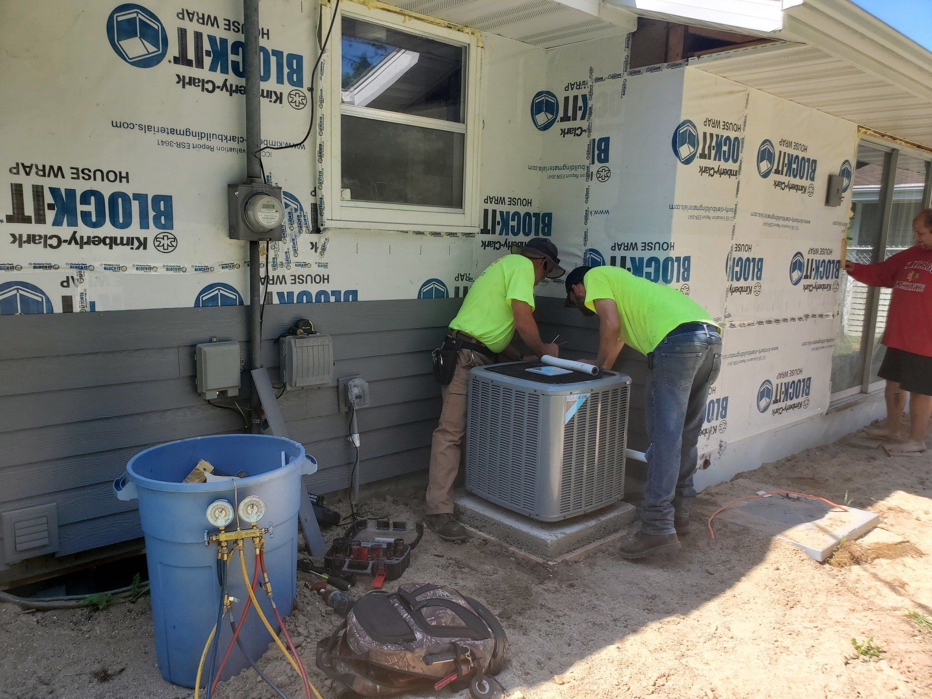 Two workers in neon shirts inspecting an outdoor air conditioner beside a house under construction.
