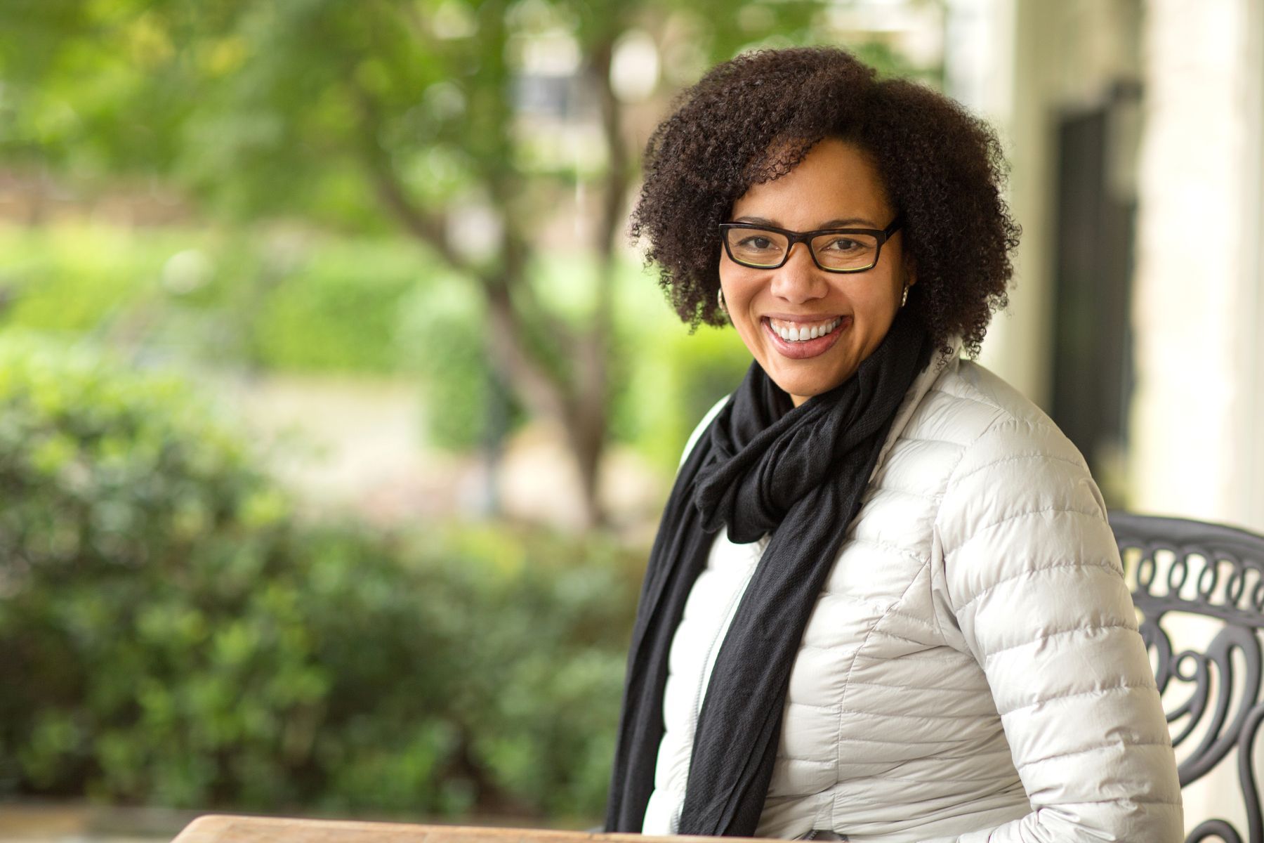 A smiling person wearing glasses, a black scarf, and a light-colored puffer jacket outdoors with a blurred green background.