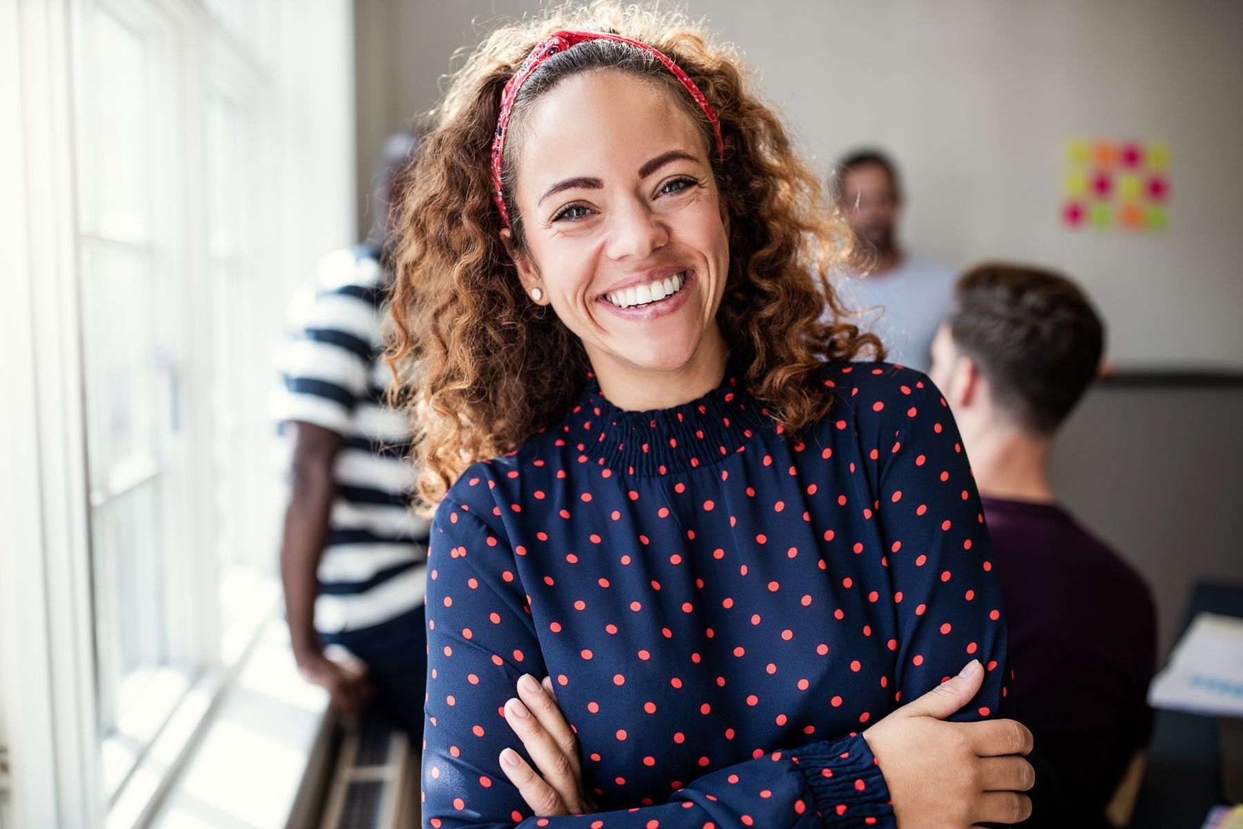 A smiling person with curly hair and a patterned top poses with arms crossed in a brightly lit office setting.
