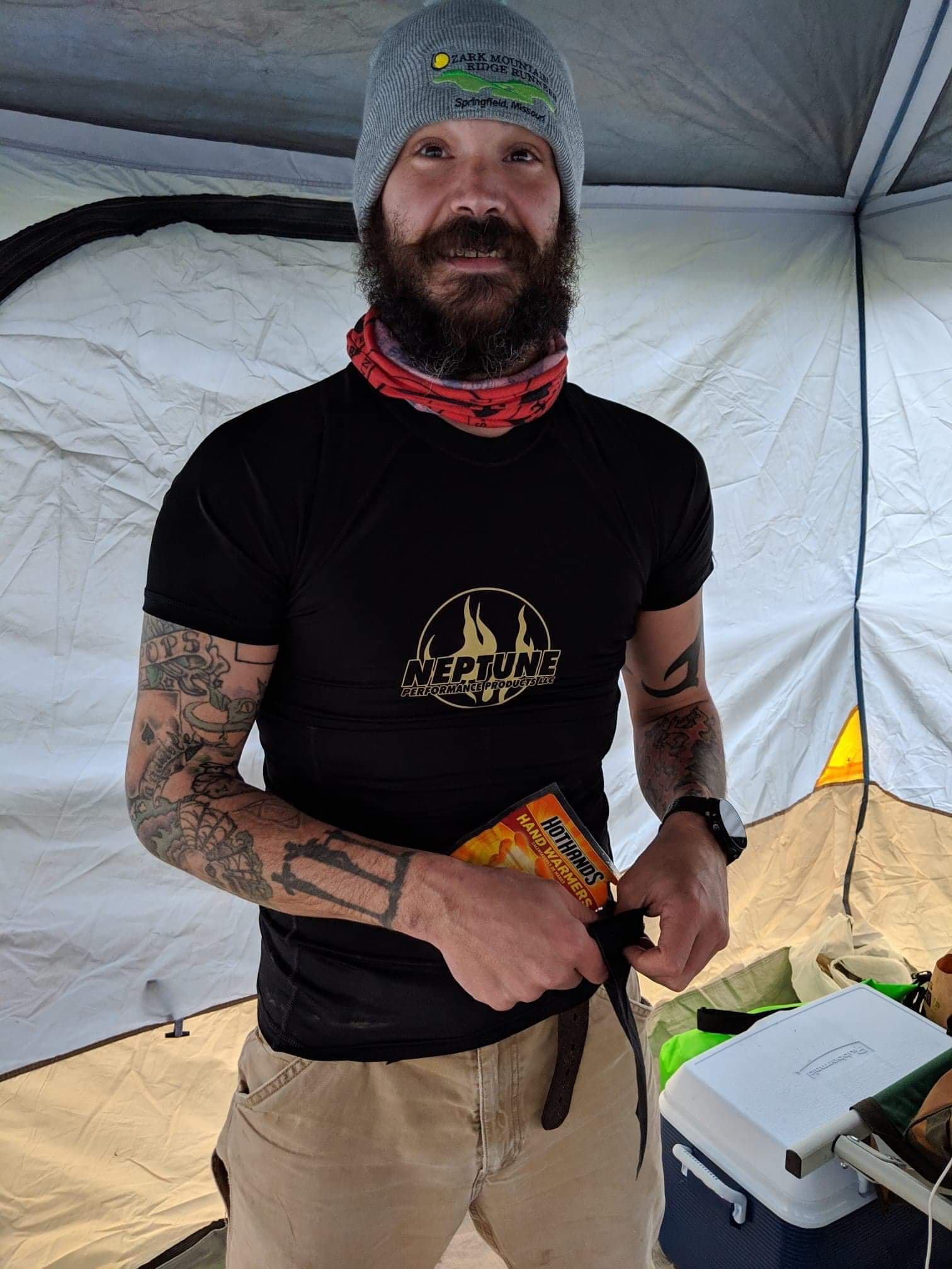 A man with a beard and tattoos is standing in front of a tent.