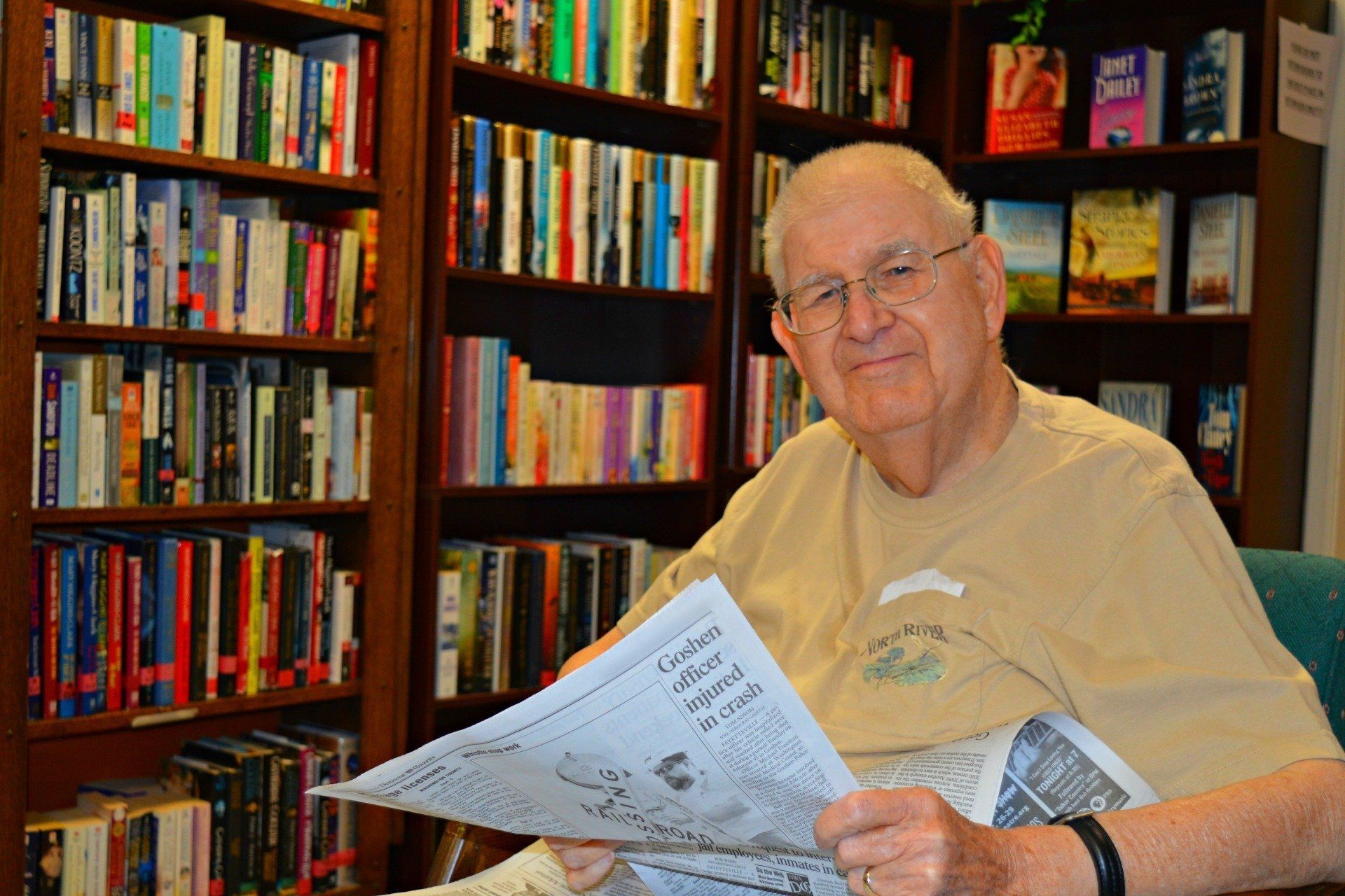 Man in glasses reading newspaper in front of bookshelves.