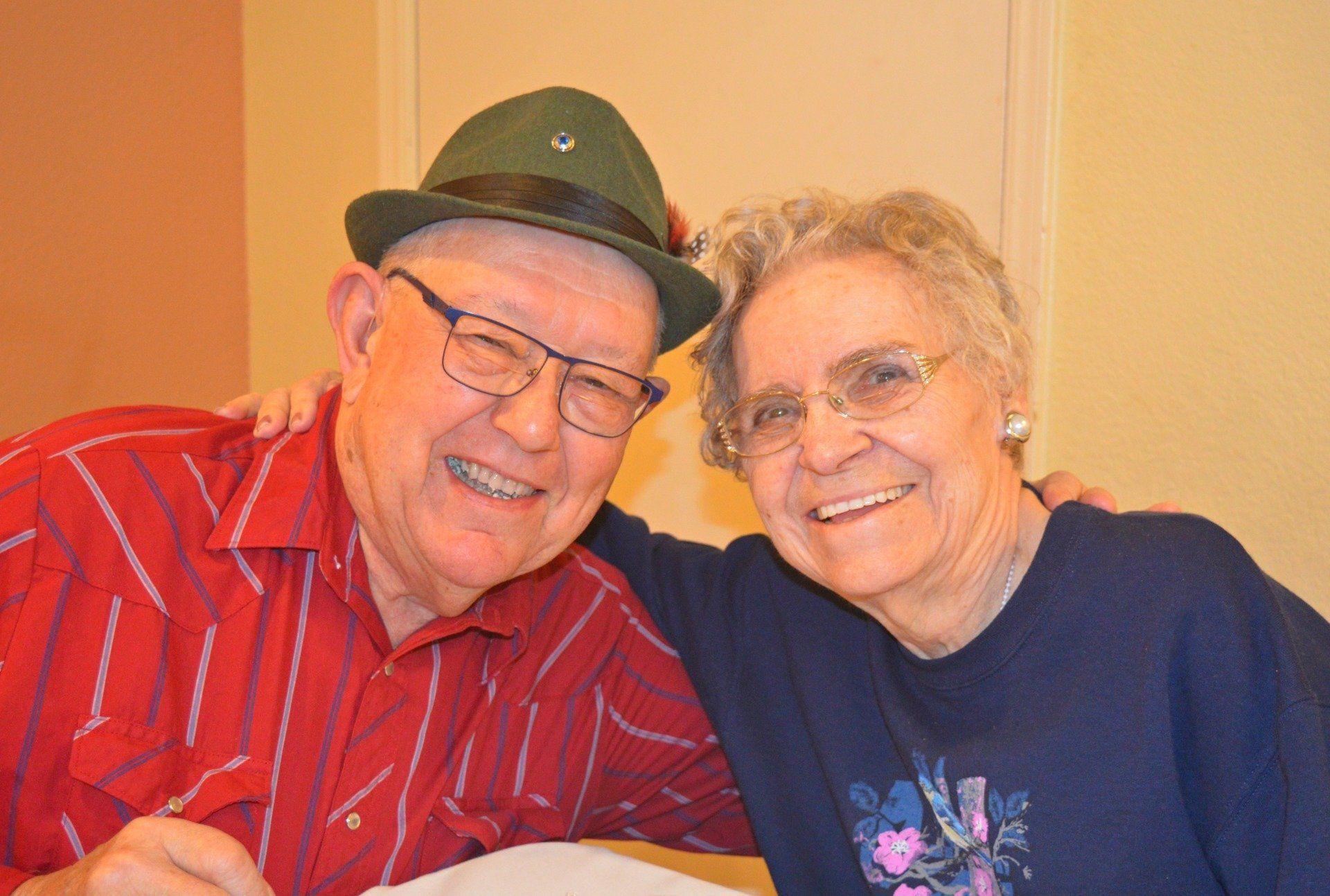Smiling couple, man in red shirt and hat, woman in blue sweater, arms around each other.