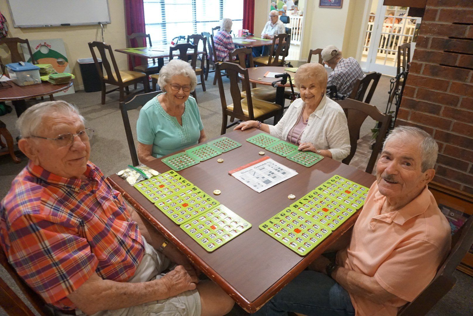 Four people playing bingo at a table indoors. Green bingo cards, smiles, and an inviting setting are present.