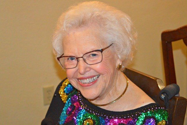 Smiling woman with glasses, white hair, wearing a colorful sequined top and a gold necklace.