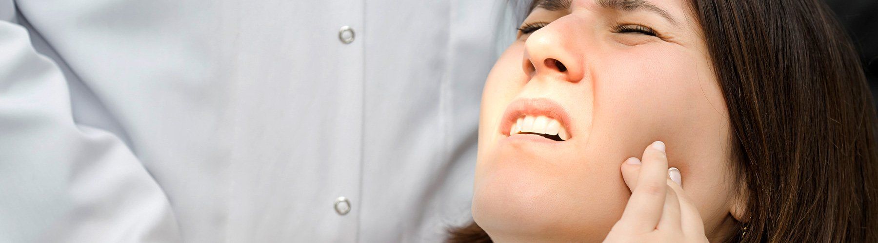 Woman in dental chair, experiencing dental pain, holding cheek while dentist in white coat stands nearby.