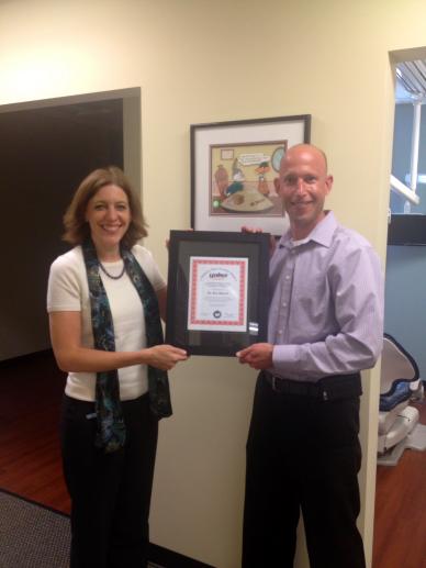 Two people, a woman and a man, holding an award in a hallway. The woman smiles, while the man grins. A framed picture hangs behind them.
