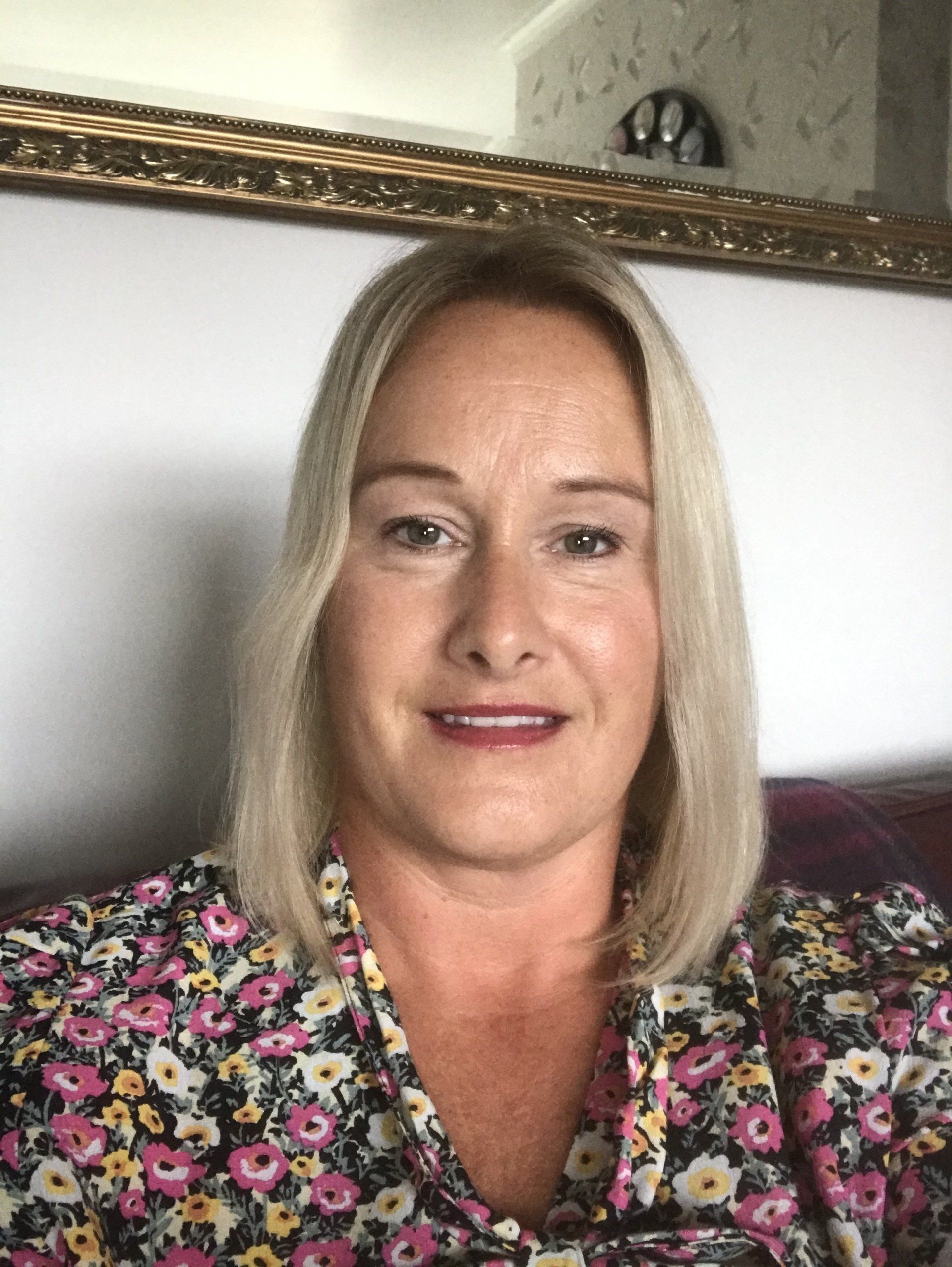 Woman with blonde bob in floral blouse, seated indoors against a light wall and framed mirror.