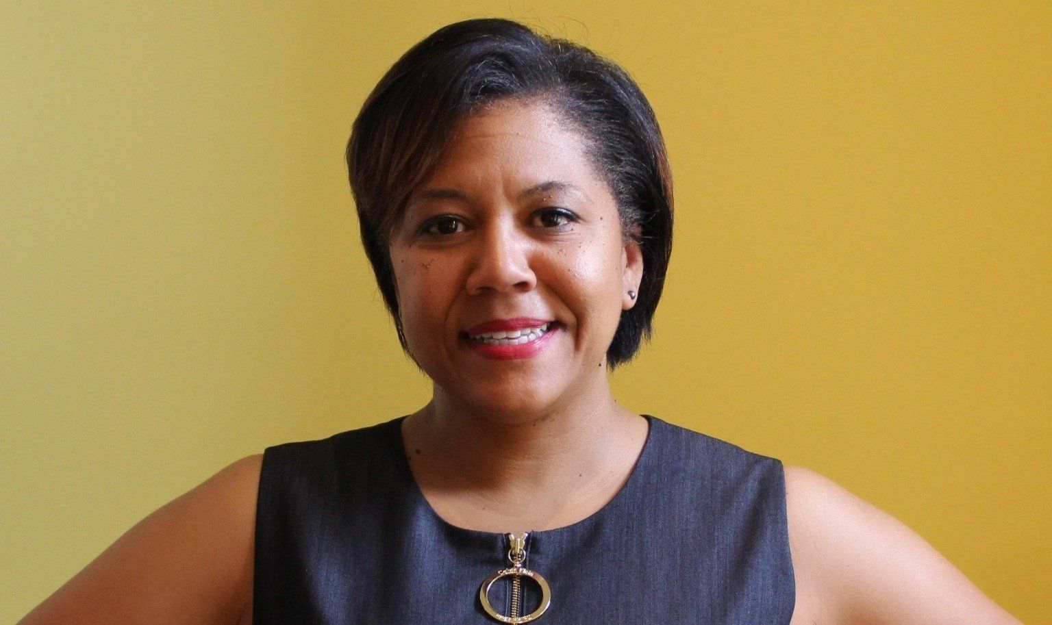 Smiling Black woman with short hair wearing a gray sleeveless top, arms akimbo, standing in front of a yellow wall.