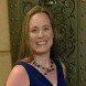 Woman in a blue dress smiles; dark necklace, long blonde hair, against a green background.