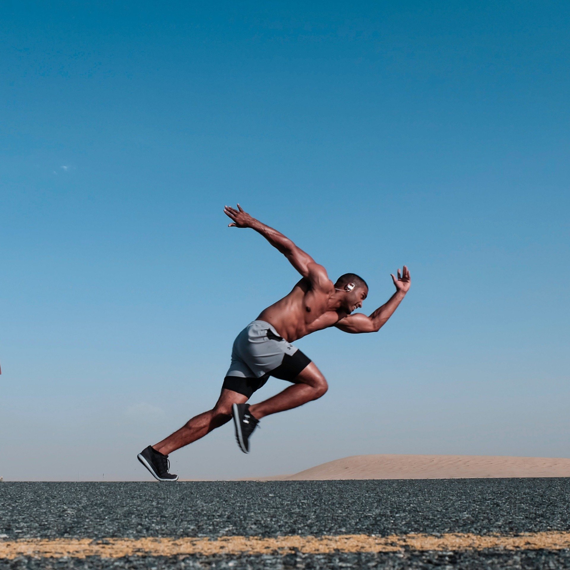 Man sprinting outdoors, shirtless, gray shorts, asphalt road, blue sky.