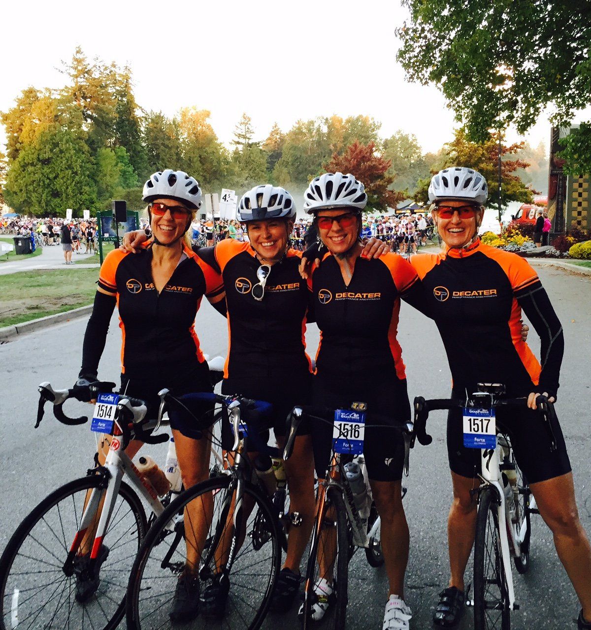 Four female cyclists in matching black and orange jerseys and helmets pose with bikes on a street, smiling.
