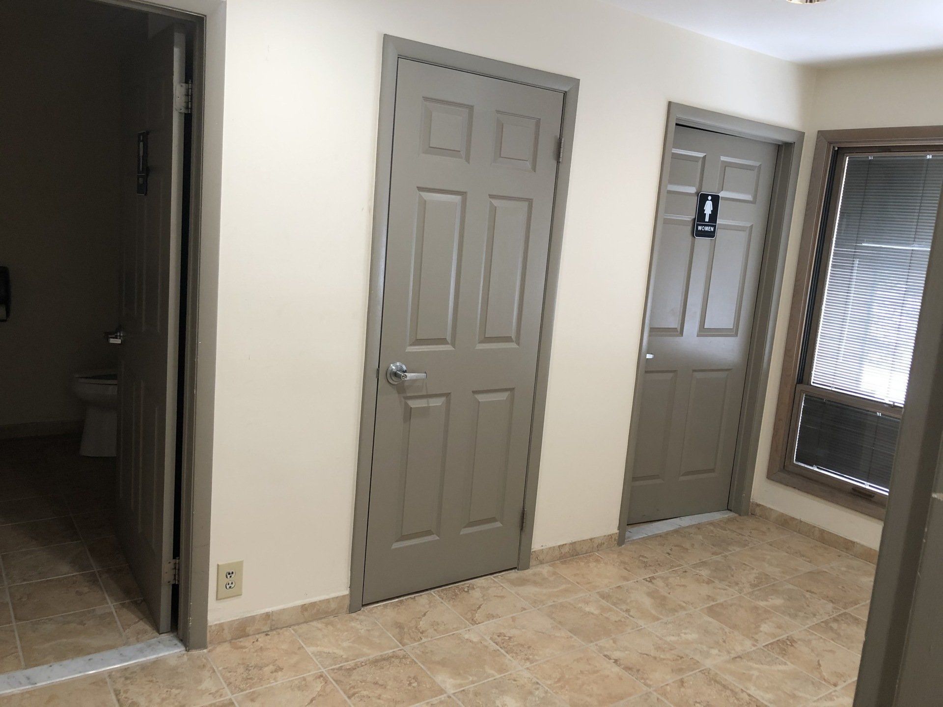Hallway with three gray doors, beige walls, and tiled floor; one door is open to a toilet.