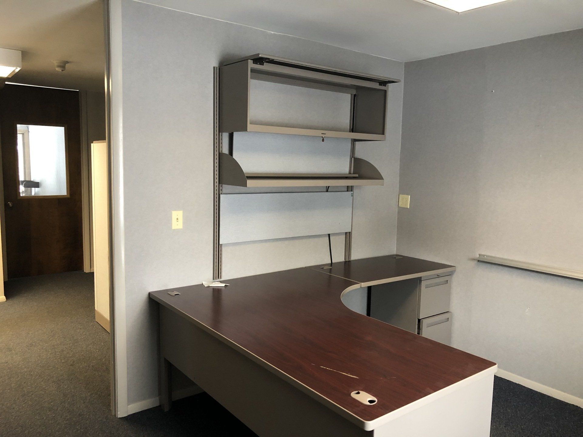 Empty office with a brown L-shaped desk, gray shelving, and a hallway.