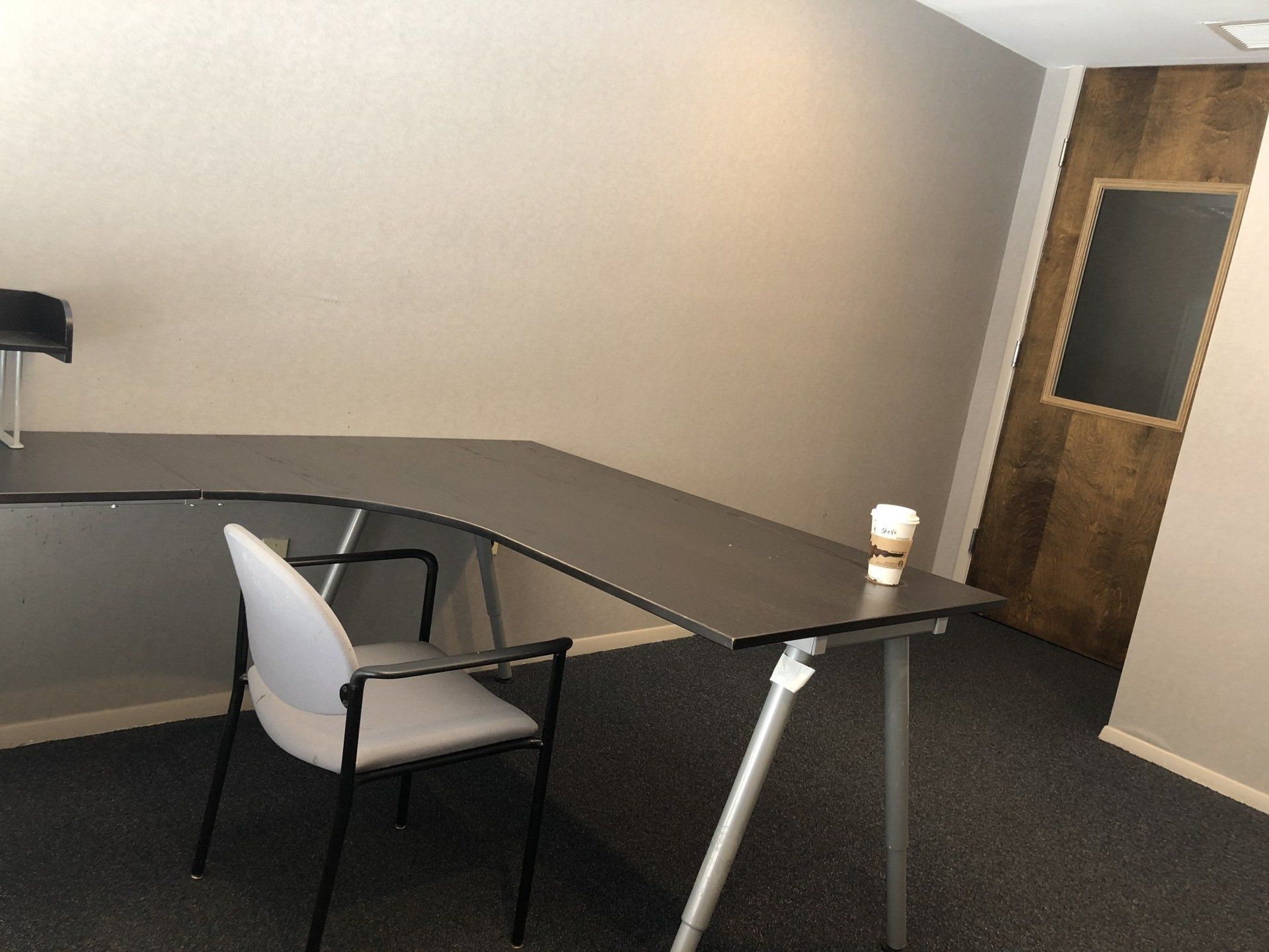 L-shaped dark desk with a white chair and a coffee cup, in a room with a wood-paneled wall and chalkboard.