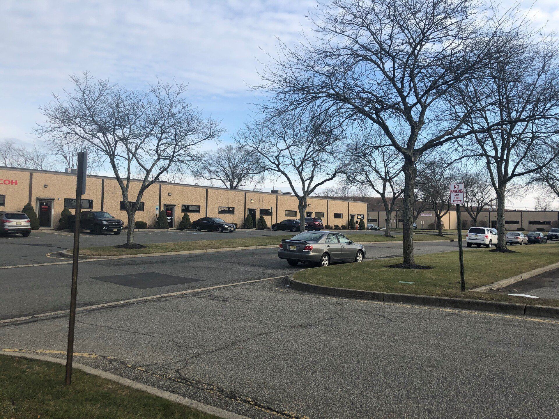 Parking lot with bare trees and a one-story beige building. Several cars are parked.