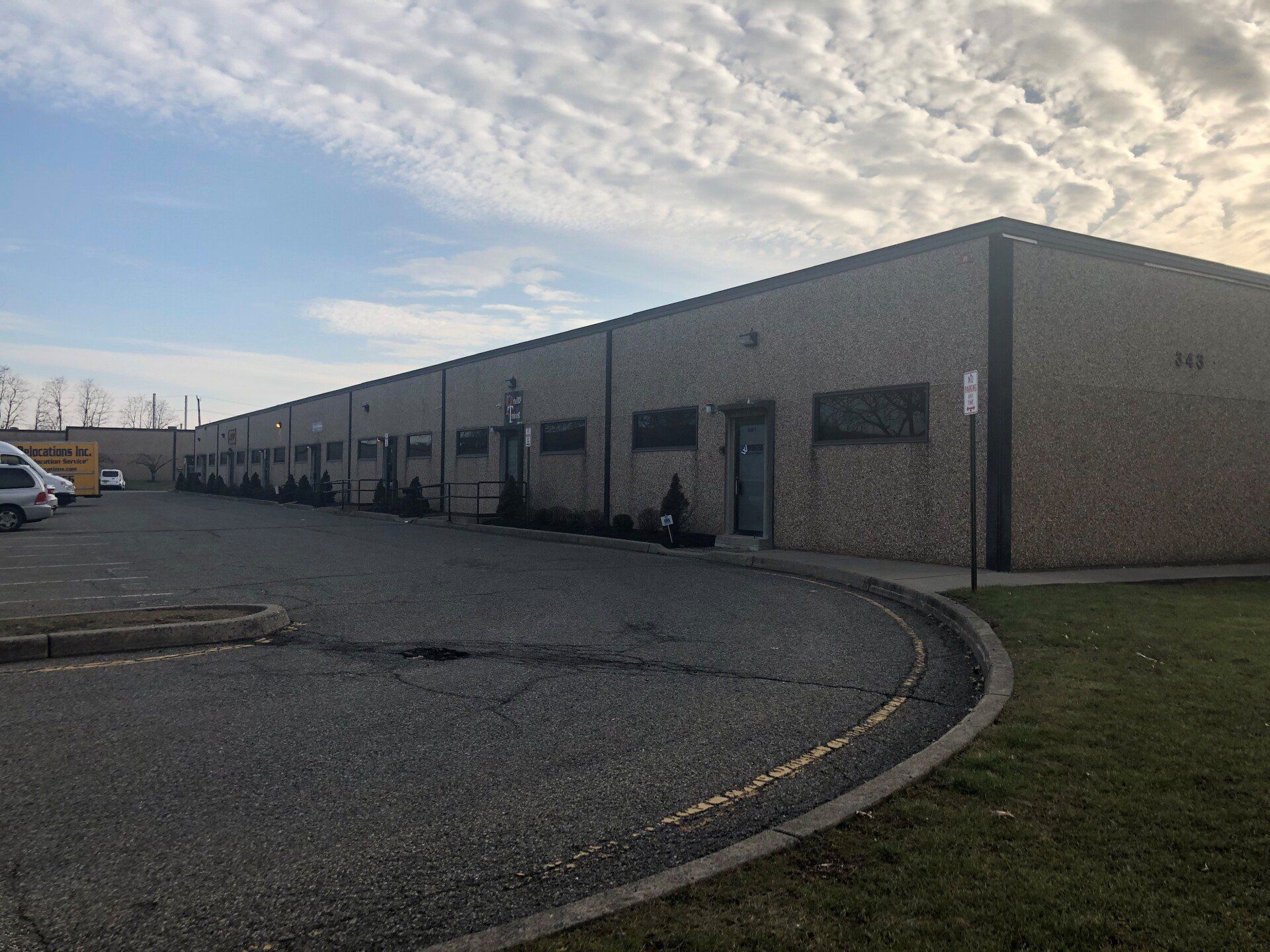 Long, single-story industrial building with multiple entry doors and windows, a cracked asphalt drive, and a grassy lawn.