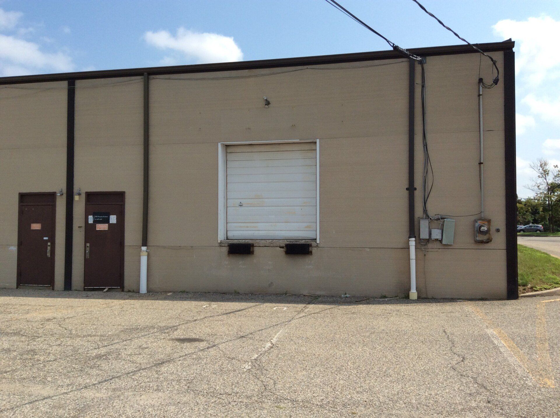 Tan building with a closed loading dock door and two brown doors on a concrete lot. Utility lines overhead.