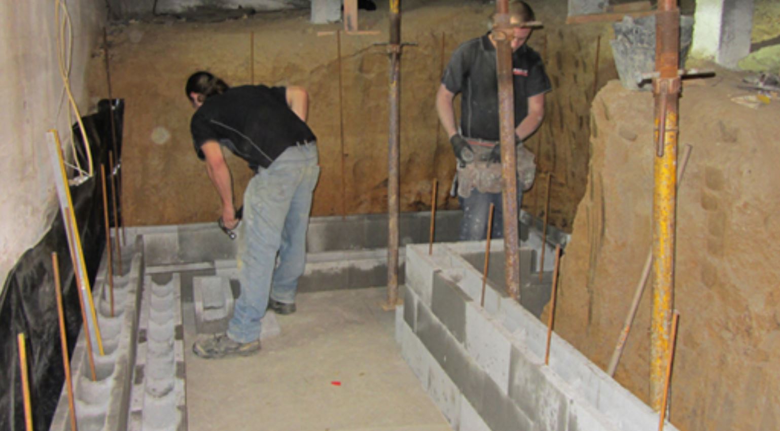 Two men are working on a concrete wall in a basement.