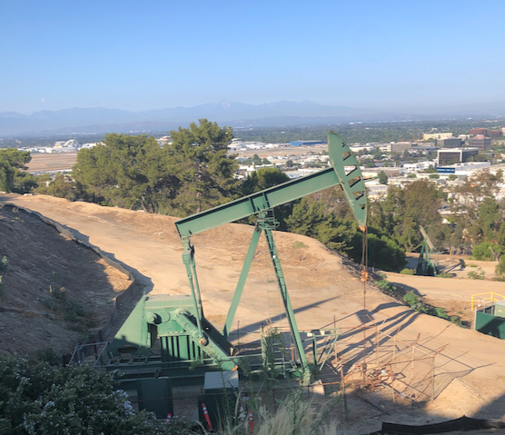 Oil pumpjack on a hillside, green against the blue sky, city and mountains in the background.