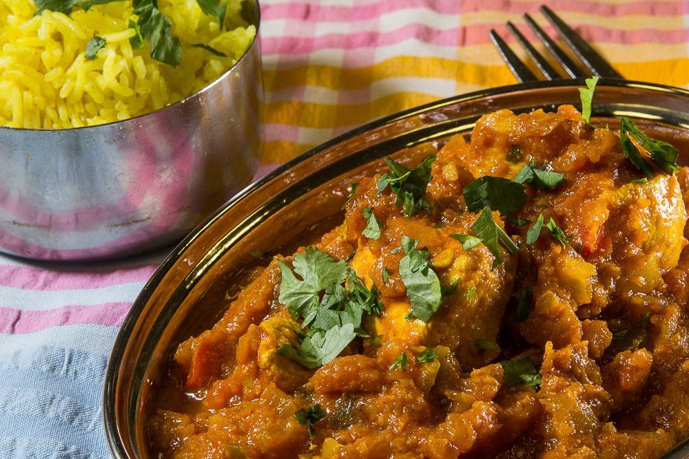 Butter chicken garnished with cilantro beside a bowl of yellow rice on a pink cloth