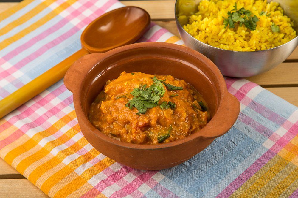Clay bowls of curry and yellow rice on a pink plaid cloth, with a wooden spoon nearby