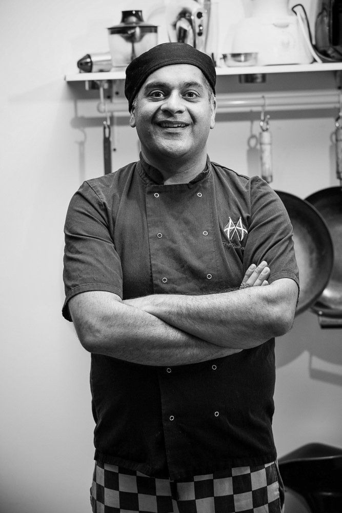 Chef standing with arms crossed in a kitchen, wearing a dark uniform and checkered apron, smiling.