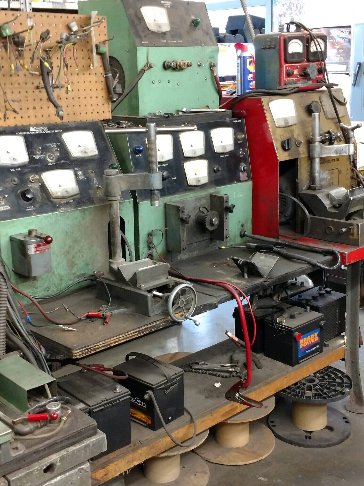 Cluttered workshop bench with vintage metal machines, gauges, cables, and batteries in red, green, and gray.
