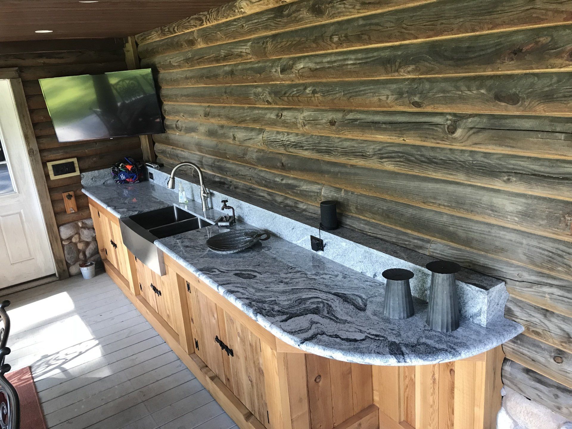 Rustic outdoor kitchen with long granite countertop, sink, stovetop, and wooden cabinets on a log wall