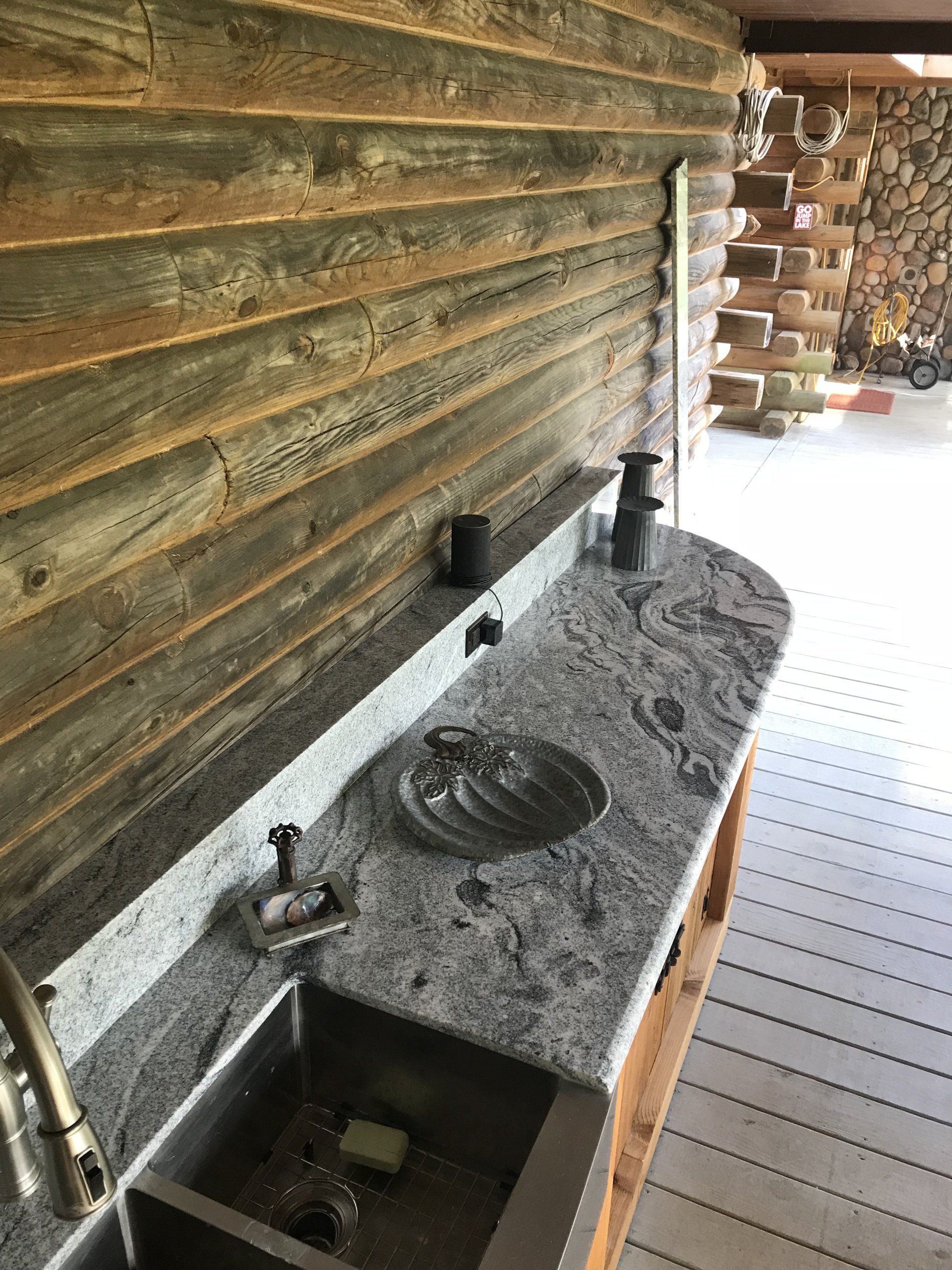 Stone sink with faucets and a soap basin against a wood wall in a rustic bathroom.
