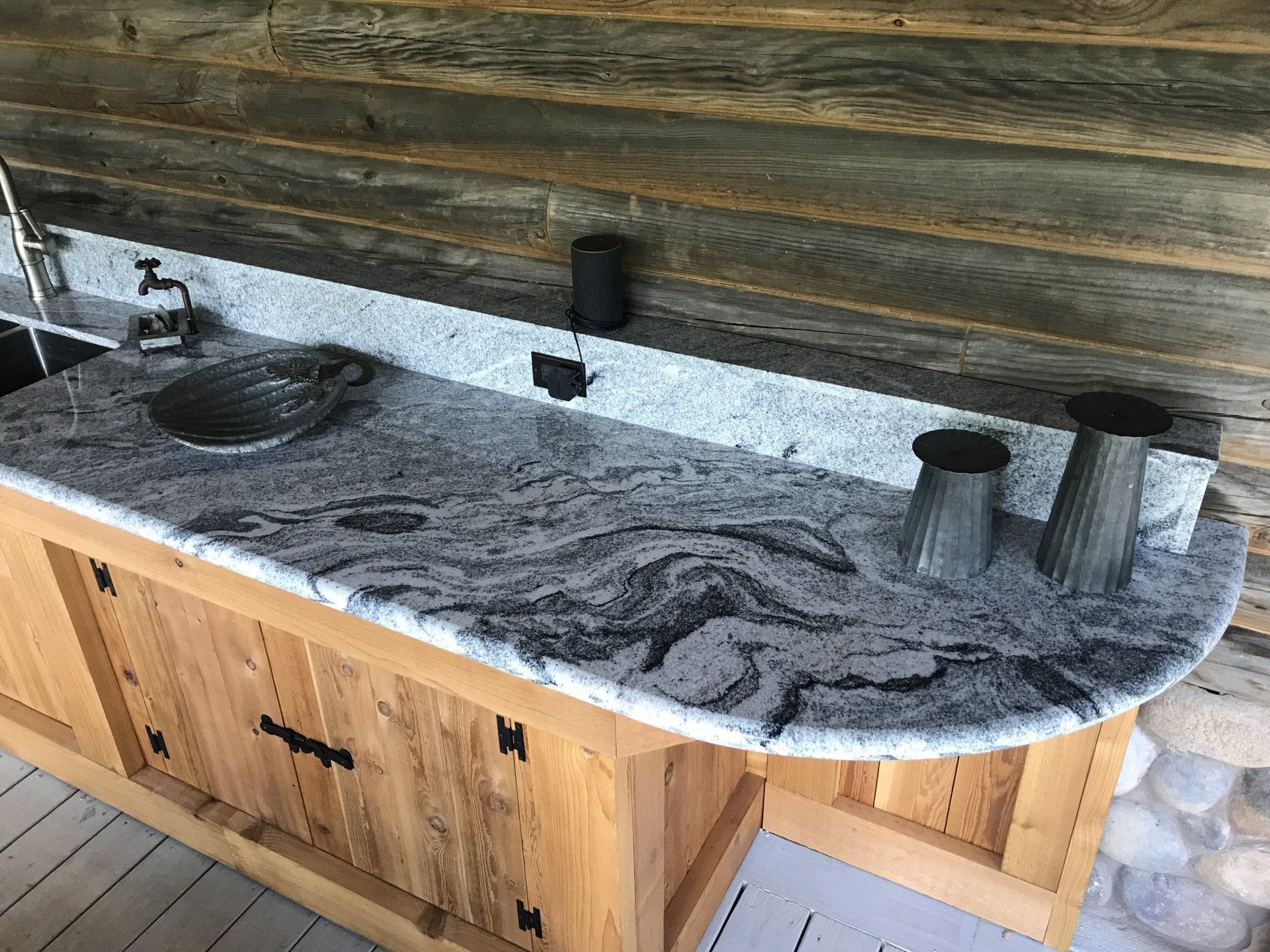 Granite outdoor kitchen countertop with sink, black faucet, and two ribbed metal cups against wood siding