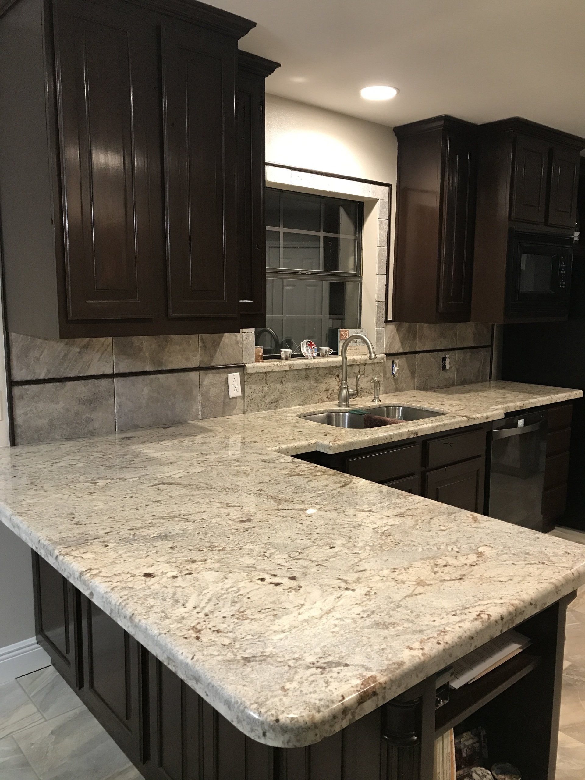 Dark wood kitchen cabinets with a light granite island and countertops around a sink and window.