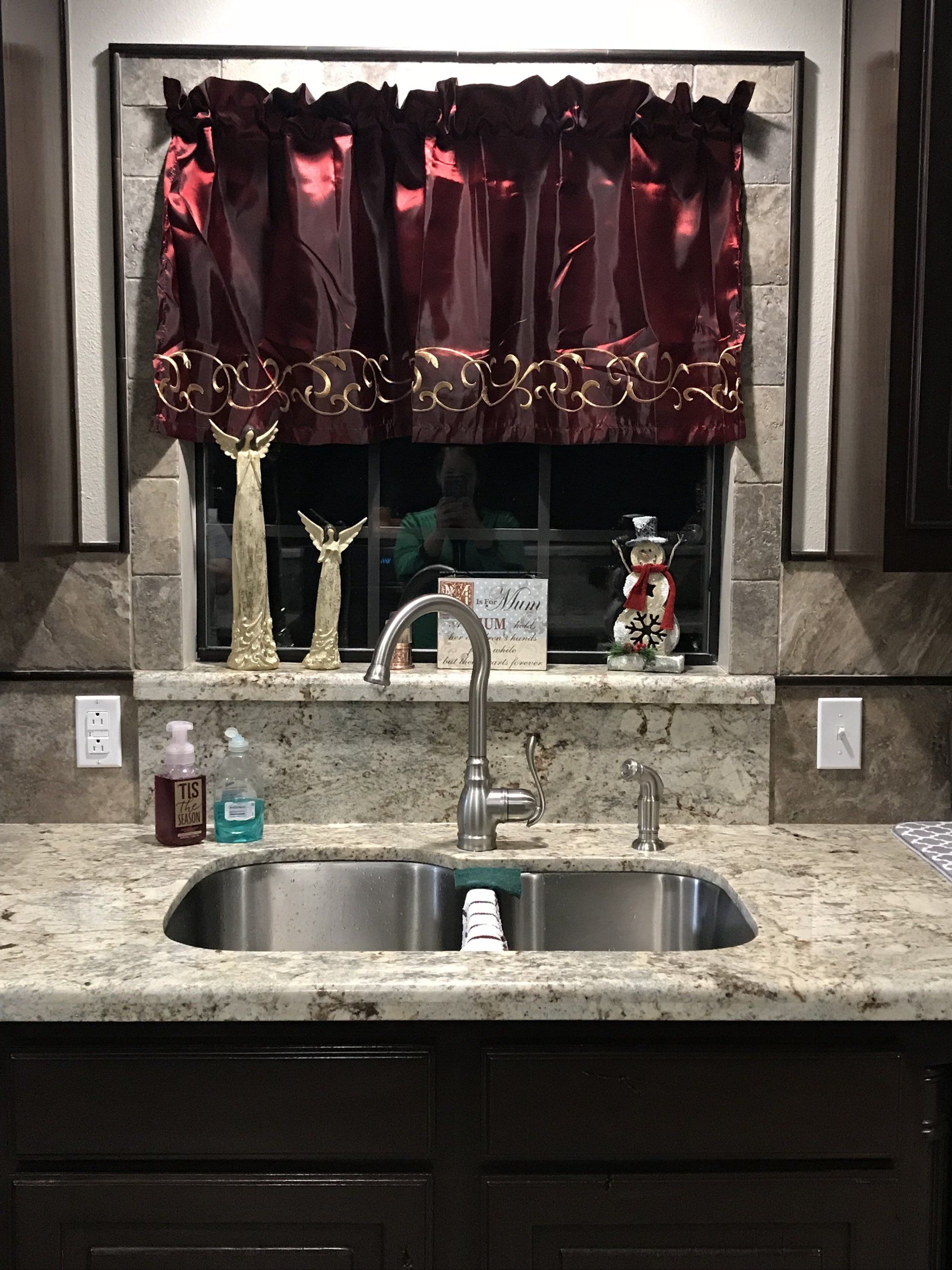 Kitchen sink beneath a window with burgundy curtains and granite countertop, showing soap and decor.