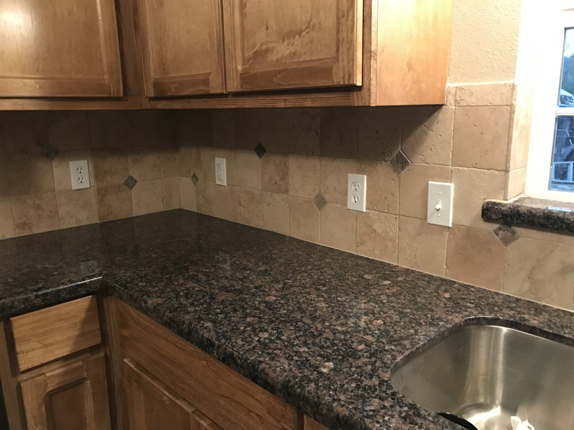 Kitchen corner with dark granite countertop, wood cabinets, beige tile backsplash, and stainless steel sink