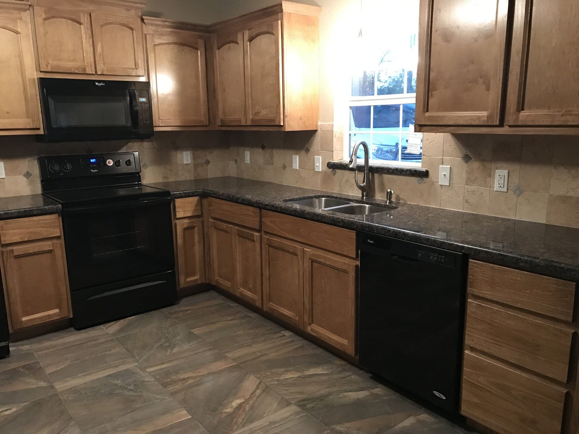 Empty kitchen with wood cabinets, black appliances, dark countertops, and a window over the sink.