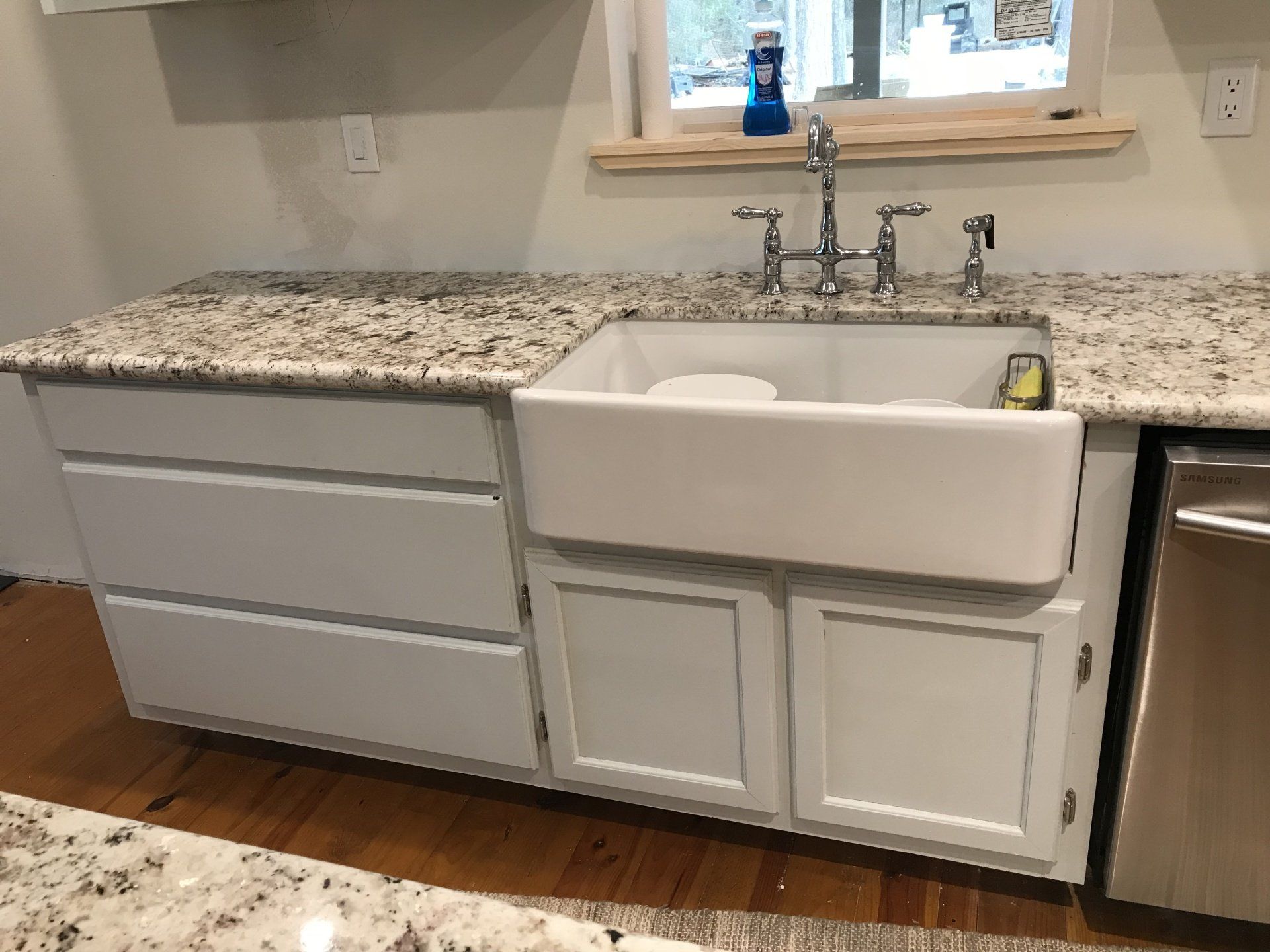 Kitchen sink with white farmhouse basin, gray cabinets, and speckled granite countertop under a window