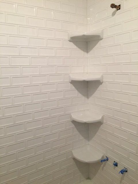 White tiled bathroom corner with staggered corner shelves and two blue-handled shower controls.