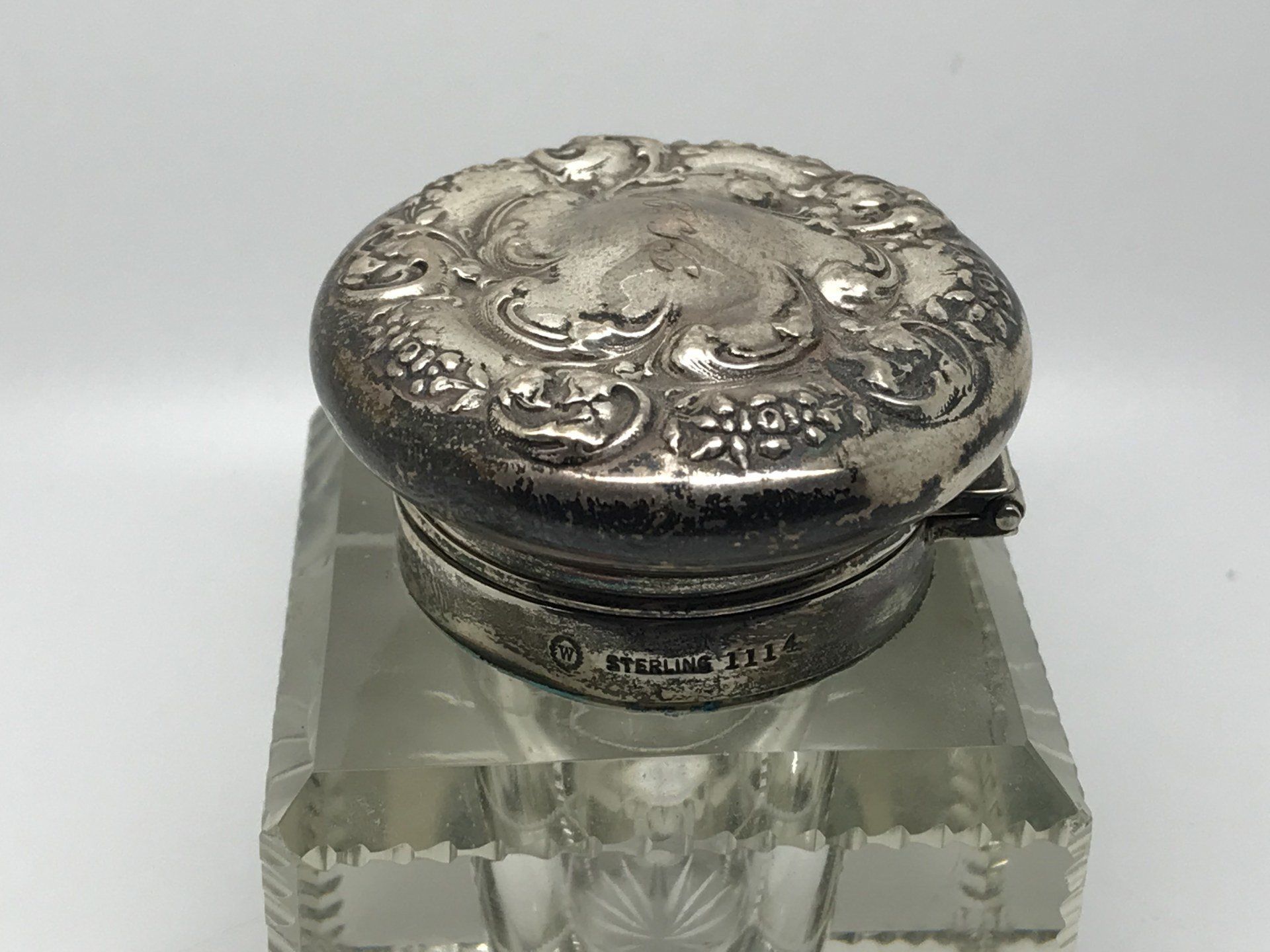 Silver-topped antique inkwell with ornate lid, set on a faceted glass base.