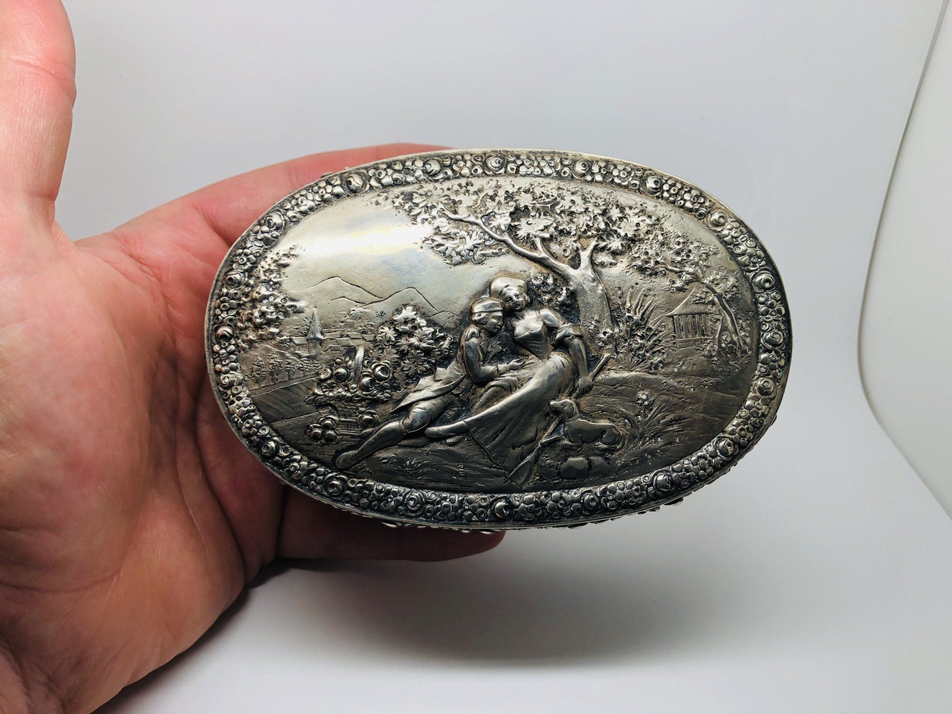 Oval silver box with relief scene of two figures among foliage.