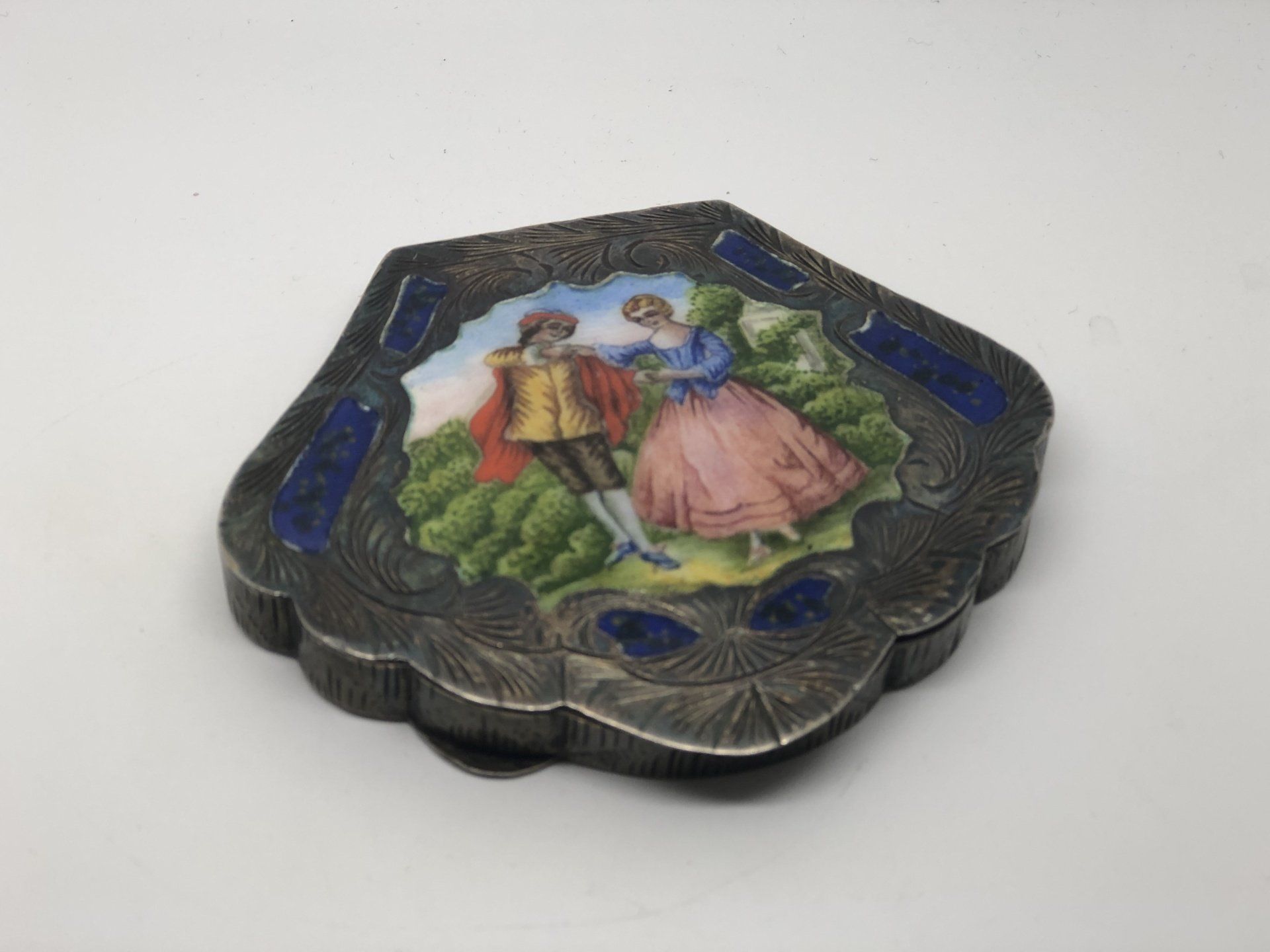 Silver box with enameled scene of two figures dancing, surrounded by blue stones.