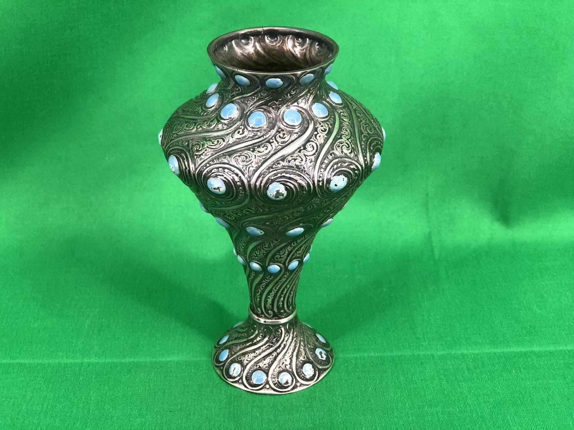 Silver vase with turquoise stones, on a green backdrop.