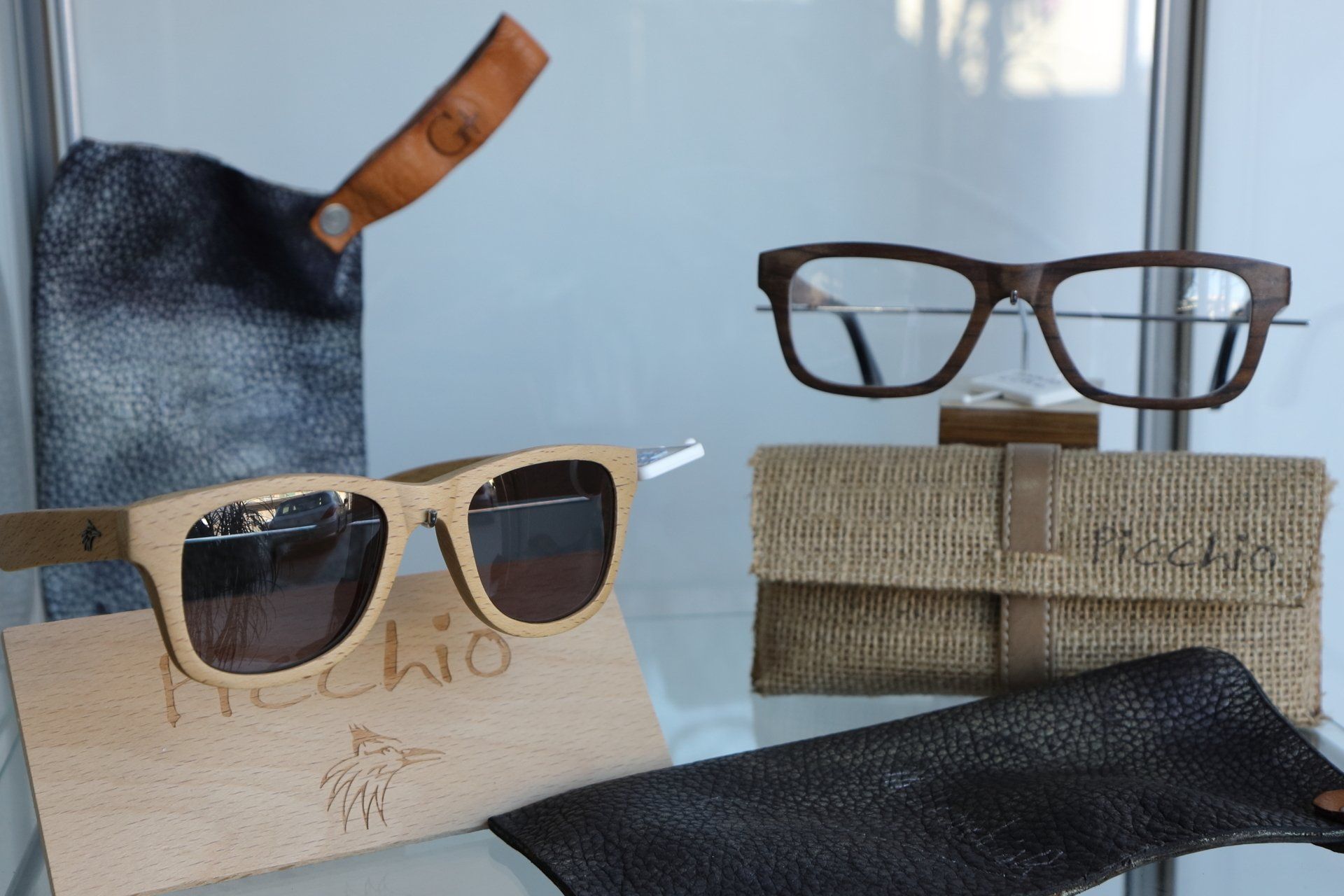 Esposizione di occhiali: montature in legno, occhiali da sole, custodie in legno e pelle, su ripiani in vetro.
