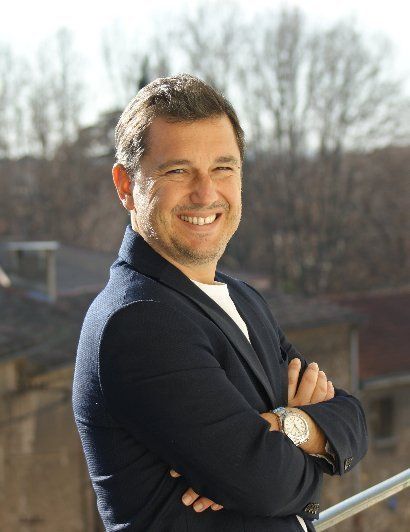 Un homme souriant, vêtu d'un blazer bleu marine et les bras croisés, se tient à l'extérieur, sur un balcon, au soleil.