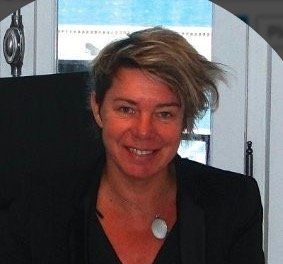 Personne souriante, vêtue d'un blazer noir et ayant les cheveux courts, assise à l'intérieur sur un fond clair.
