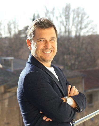 Un homme souriant, vêtu d'un blazer sombre et les bras croisés, se tient à l'extérieur en plein soleil.