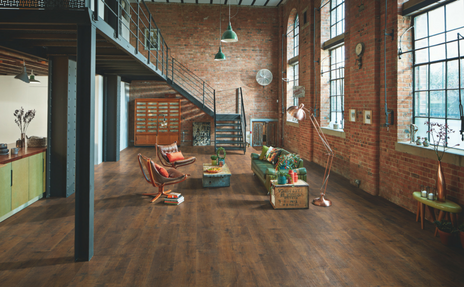 Loft-style office with brick walls, wooden floor, staircase, desks, chairs, and large windows.