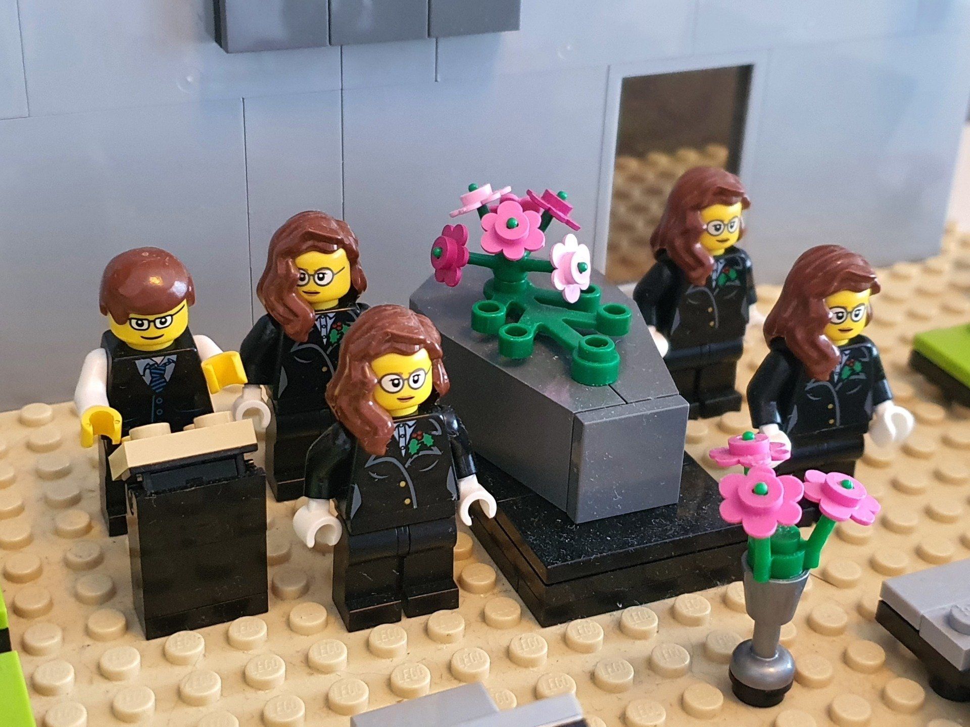 Lego-figuurtjes in galakleding staan ​​rond een grijze doodskist versierd met roze bloemen tijdens een begrafenis.
